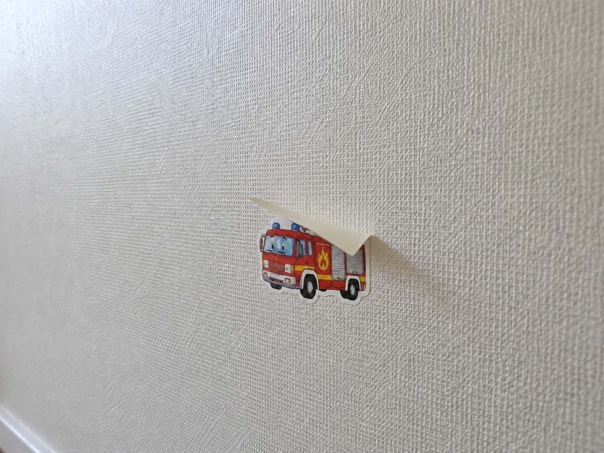 Ein kleiner, cartoonartiger Feuerwehrauto-Sticker klebt auf einer hellen, strukturierten Vinyltapete. Die obere Kante des Stickers löst sich deutlich von der Wand und hängt nach unten, wodurch ein sichtbarer Schatten entsteht.