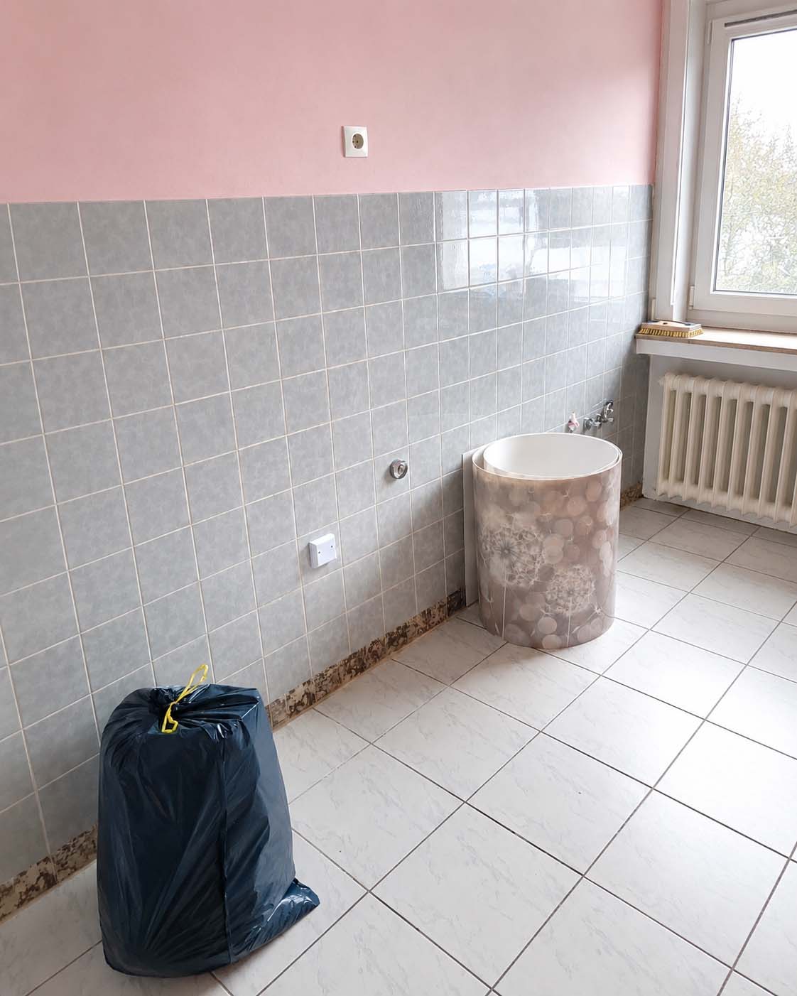 Helle K&uuml;che mit grauen Wandfliesen, rosa oberer Wand, wei&szlig;em Fliesenboden, Fenster mit Heizk&ouml;rper, Wasseranschluss und K&uuml;chenr&uuml;ckwand neben einem schwarzen M&uuml;llsack.