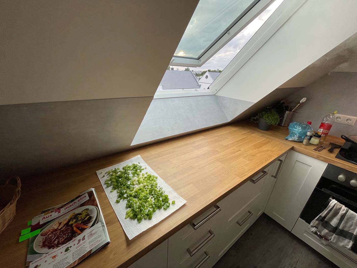 Küche mit Holzarbeitsplatte unter einem geöffneten Dachfenster; auf einem Küchentuch liegen frisch geschnittene Frühlingszwiebeln, daneben ein Kochheft, weiße Unterschränke, Herd und Küchenutensilien.